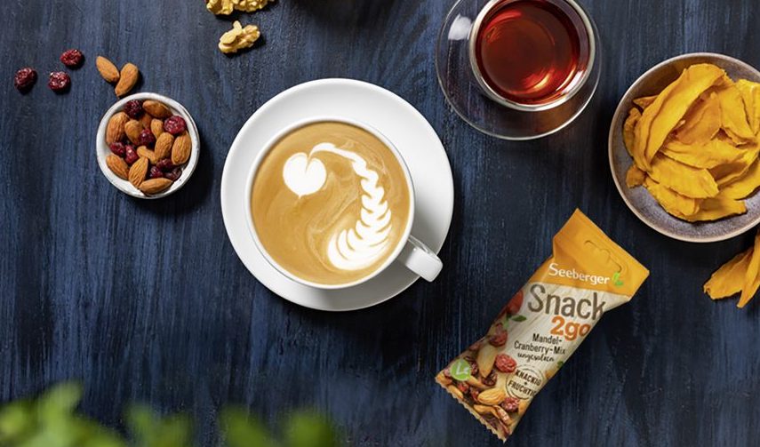 Kaffee mit Latte Art, Snack to go und Schale mit Nüsse und Früchten auf blauem Holzhintergrund aus der Vogelperspektive